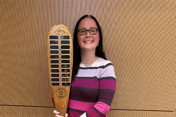 A woman with brown hair wearing glasses and pink and purple striped shirt holding a row boat oar with plaques on it.