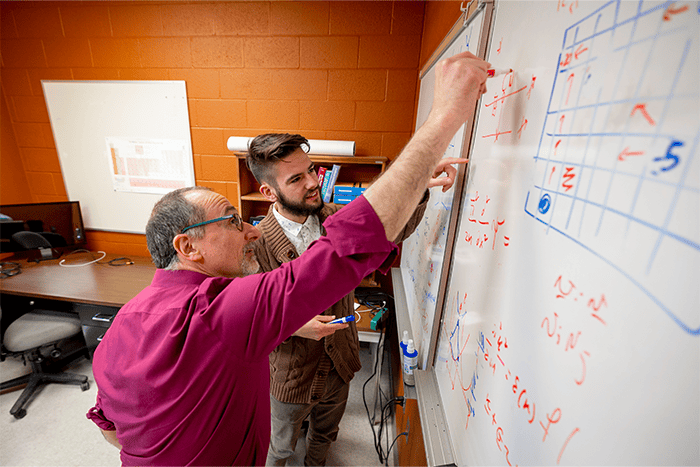 Man with glasses wearing a maroon shirt and man with beard wearing brown sweater writing on whiteboard.