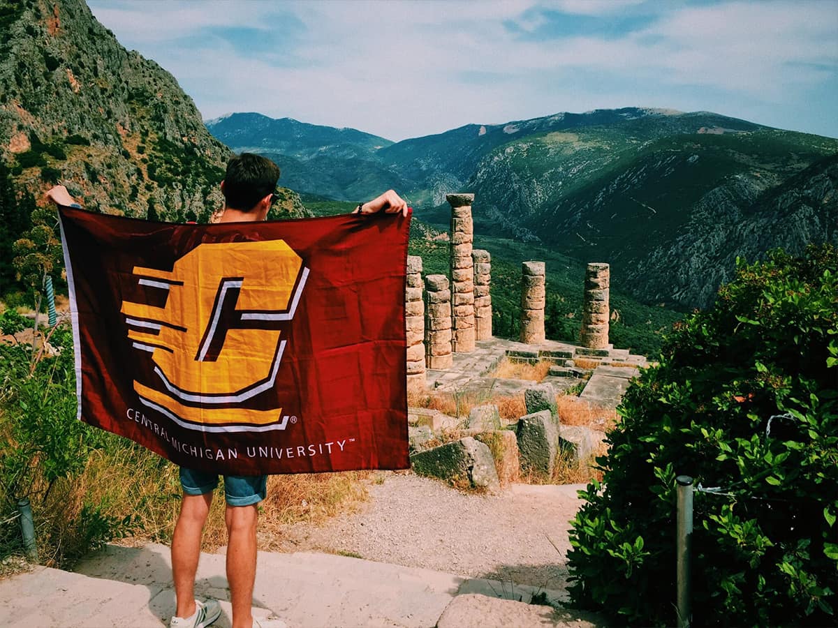 A young man stands in front of ancient Greek ruins while holding a CMU flag.