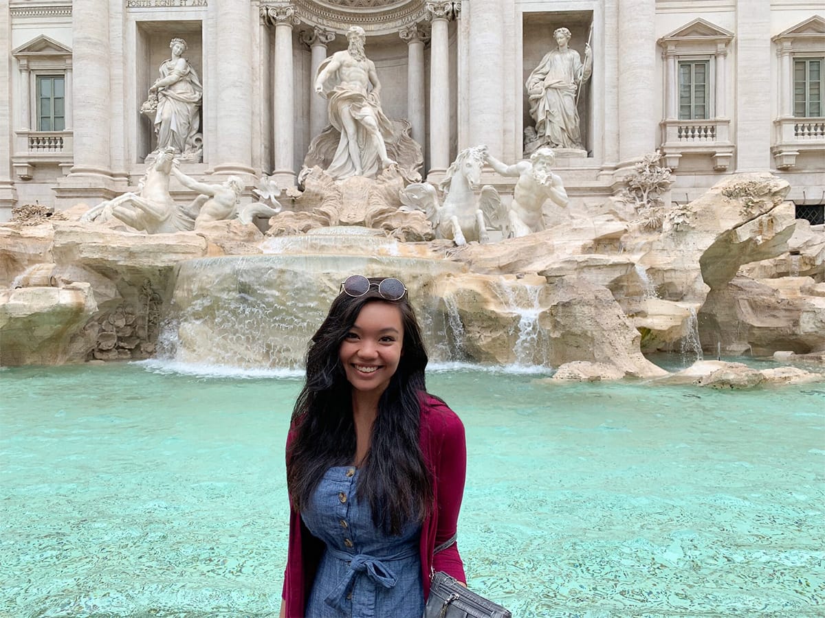 A woman in a denim dress and sweater poses in front of a fountain in Italy.