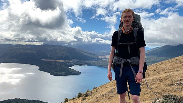 Student with a backpack standing on a mountain overlooking a lake and other mountain views in New Zealand. 