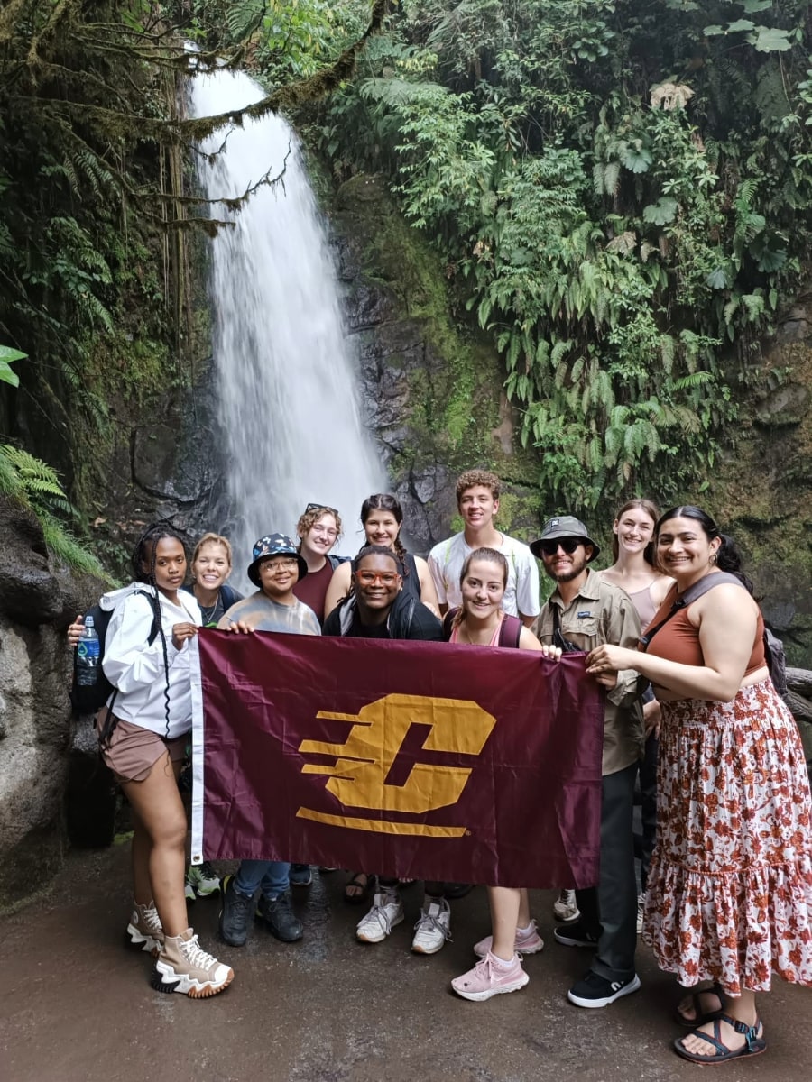 Study Abroad students holding a Central flag, standing in front of a waterfall surrounded by lush greenery.