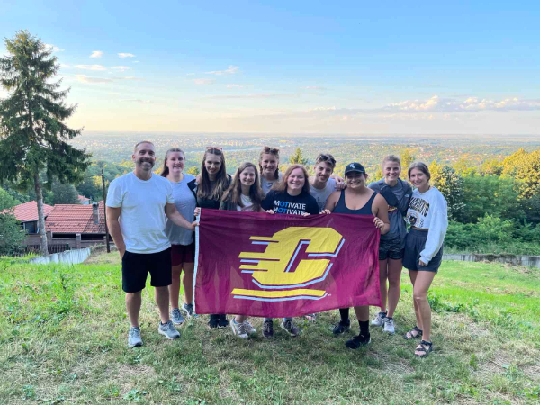 Study abroad faculty and students holding up a central flag, standing on a grassy terrain with a backdrop of greenery, trees, and buildings under a clear sky.