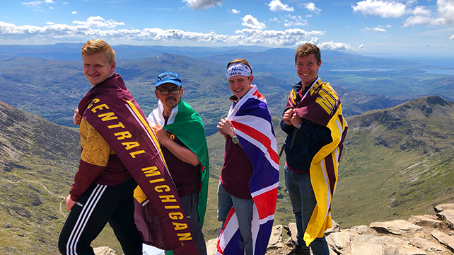 Four men standing on a mountain with large flags wrapped around their shoulders.