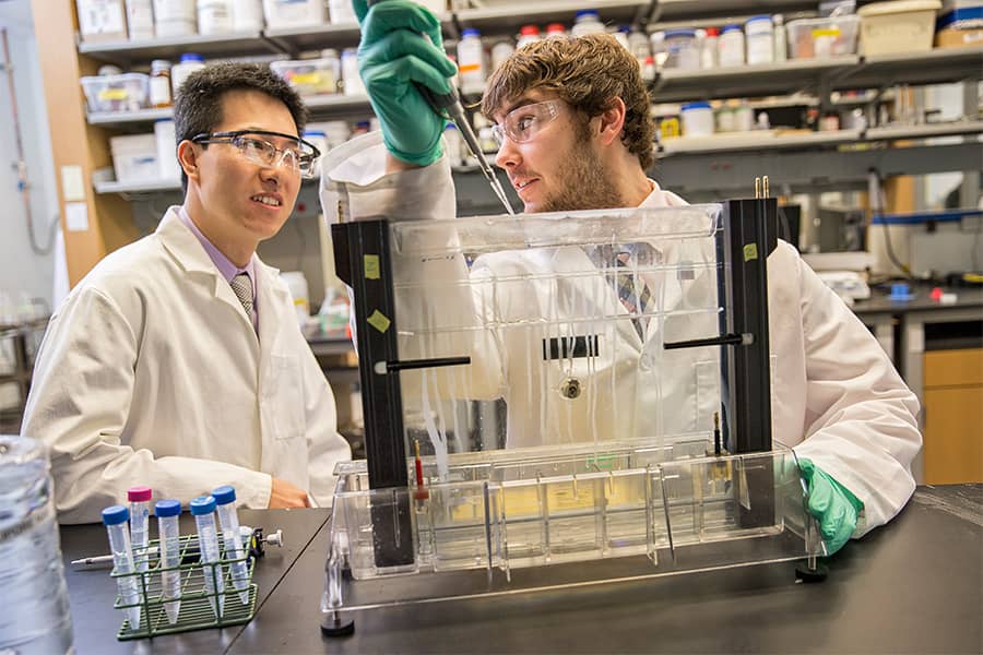 Two students wearing lab coats and goggles using lab equipment.