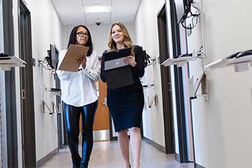 Two health administration students dressed professionally stand side-by-side holding clipboards and papers and walk down the hallway of the Interprofessional Education and Practice Center.