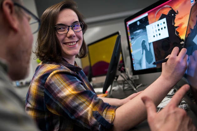 A student working on a computer and smiling while looking to the side at someone else.