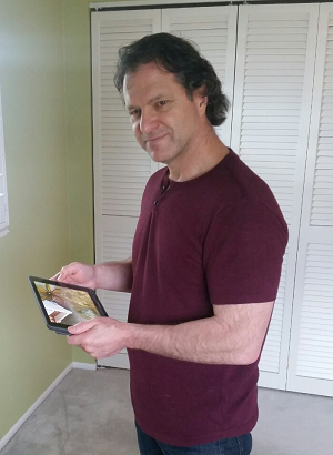 George Thayer wears a maroon t-shirt while standing and holding a tablet.