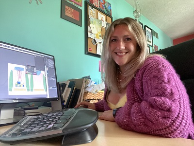 Katelyn Haas working on clothes designs on the computer while smiling at the camera.