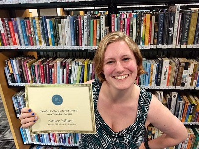 Aimee Miller wearing a black dress is while holding an award and smiling at the camera.