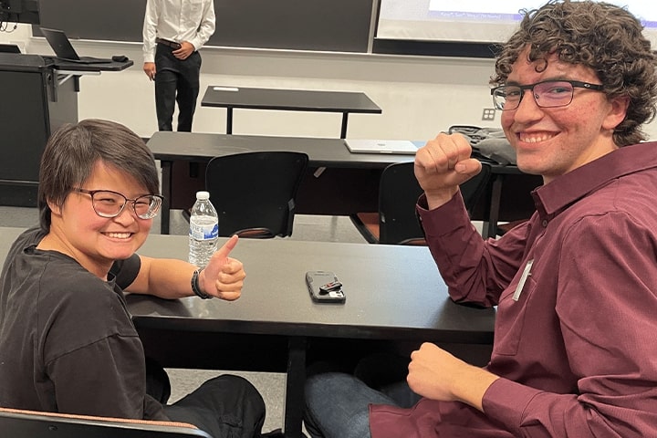 Two students with glasses smiling for a photo in a classroom