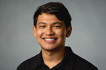 A man in a black shirt smiling for a photo in front of a gray background.