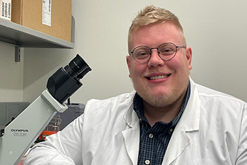 A man with short blonde hair wearing glasses and a lab coat is next to a microscope.