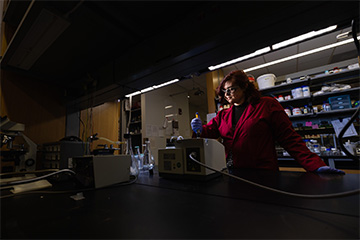 A woman in a red lab coat and safety goggles working in a dark lab.