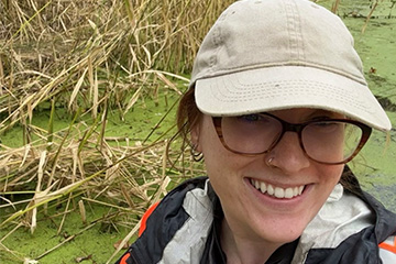A woman in a tan hat and glasses taking a selfie in a swamp.