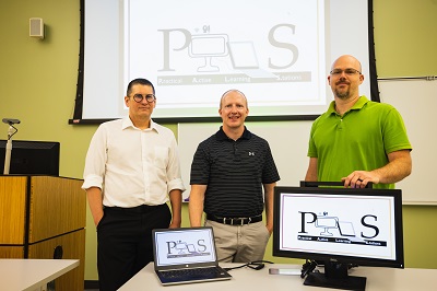 Three people standing behind two computers displaying the PALS logo.