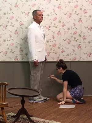 A man standing next to a wall while wearing a suit with a woman spray painting his pants.