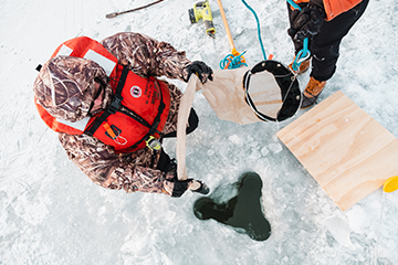 A man in a camo jacket kneeling in front of a hole in ice.
