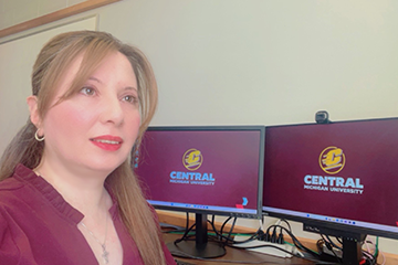 A woman in a maroon shirt in front of two computer screens