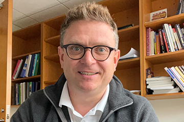 A man with glasses and a collared shirt smiling in an office.