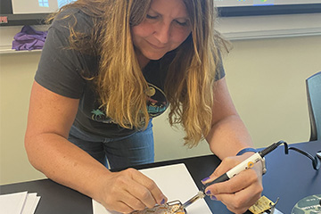 A woman in a t-shirt soldering a piece of technology.