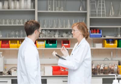 Two students wearing lab coats and safety glasses standing and talking.