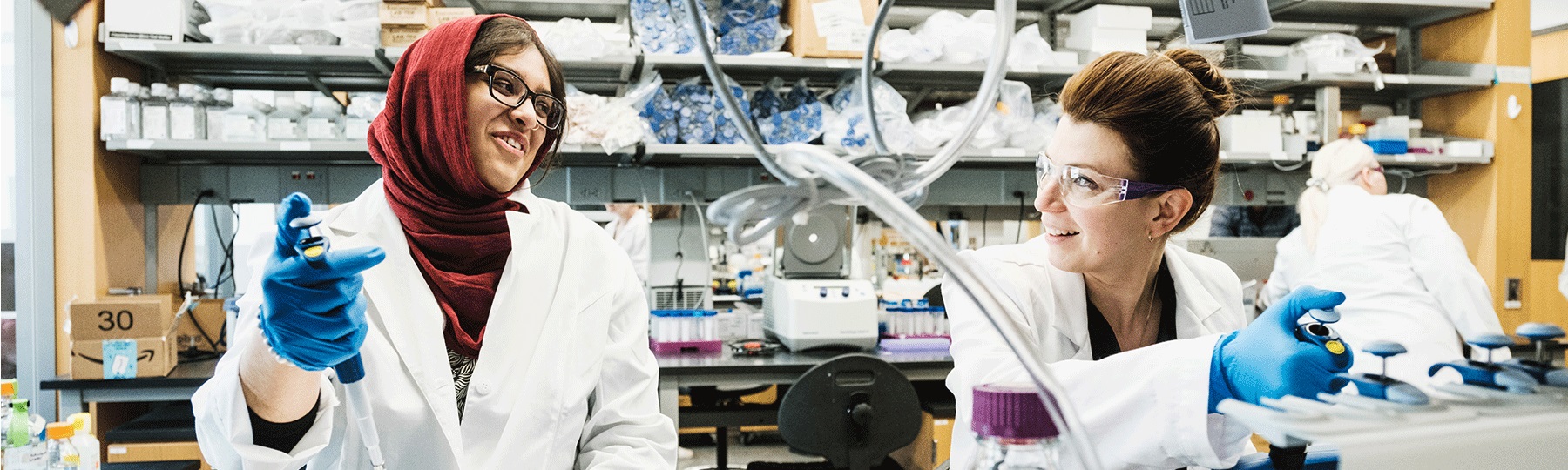 Two female students in white lab coats work with pipettes in a sepsis lab on campus.