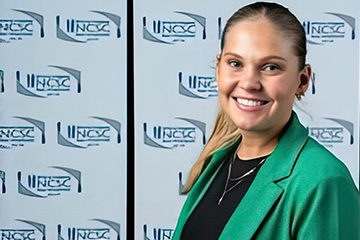 Keirra Ryan stands in front of a logo backdrop with a ponytail dressed in a green blazer, black shirt and necklace.