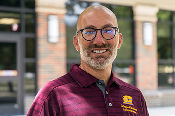Sal Buccellato smiles outside a campus building, wearing glasses and a maroon Central Michigan University College of Business Administration polo.