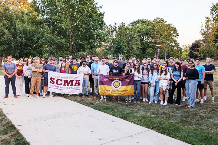 Fred Lawrence (far left in blue polo and dark blue pants) stands with a large group of Supply Chain Management Association students on the Warriner Mall. The students are holding SCMA flags and posing as a group.