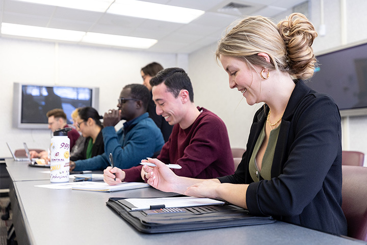 Students sit in a classroom during a lecture. In the foreground a young woman with blond hair in a bun laughs and looks at her padfolio. A faculty member can be seen chatting with students in the background.