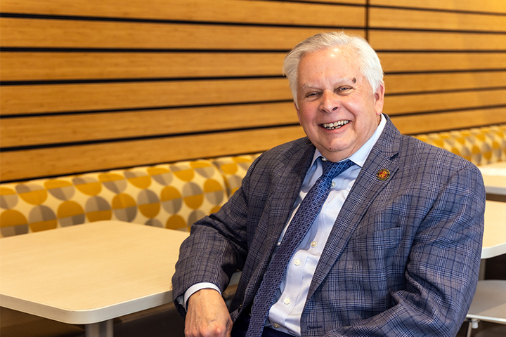 Thomas Anthony sits at a booth table in Grawn Hall. He's wearing a dark blue plaid jacket with a white shirt and blue tie. He has white hair and is smiling.