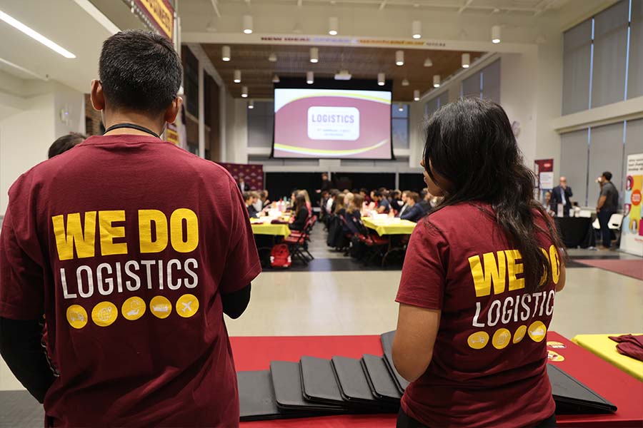 A photo from ERPsim, two students are seen standing with their backs facing the camera as they wear a maroon and gold t shirt with the words We Do Logistics. They are inside of a large conference room with a screen in the center of them.