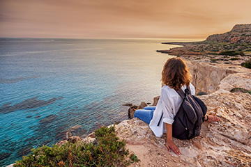 A woman in a white shirt and backpack sits on the edge of a sandstone cliff next to a large body of water. The surrounding terrain is sparse and rocky. The sky is a gentle orange and yellow and the water is calm.