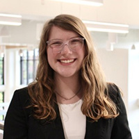 Britta Williams smiling in a professional headshot, wearing glasses and a black blazer in a bright indoor setting.