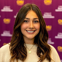 Carmella Cortes smiling in a professional headshot, wearing a light sweater with a maroon CMU logo backdrop.