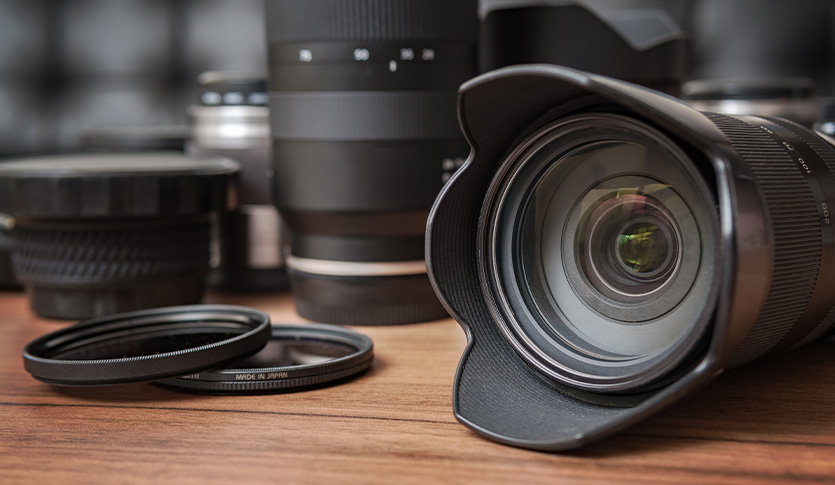 Close-up of a camera lens on a wooden table with additional lenses and filters in the background.
