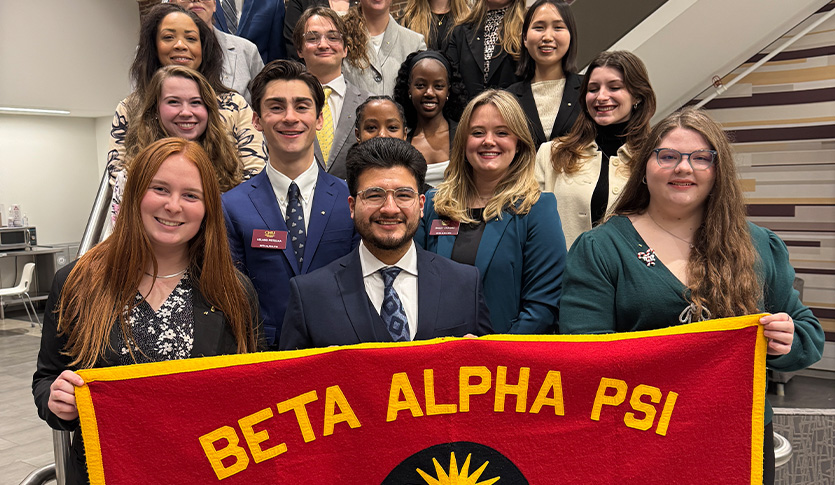 Beta Alpha Psi members pose for a group photo indoors, with students in the front row holding a large maroon and gold Beta Alpha Psi banner.