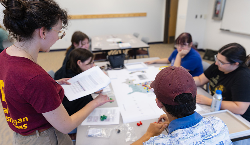 A group of students gathers around a table in a library meeting room to test a tabletop game, with one participant explaining the rules while others examine game pieces and materials.