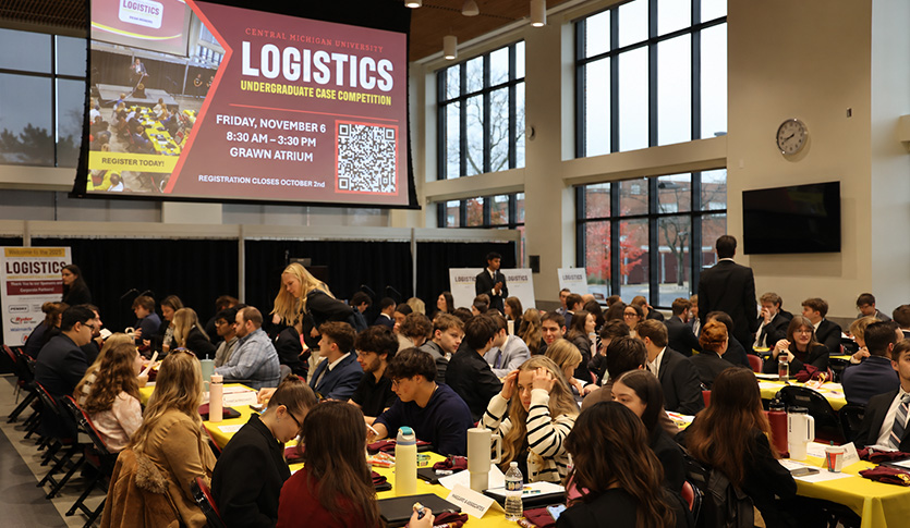 Attendees gather around tables in Grawn Atrium beneath a projected slide promoting CMU’s Logistics Undergraduate Case Competition.