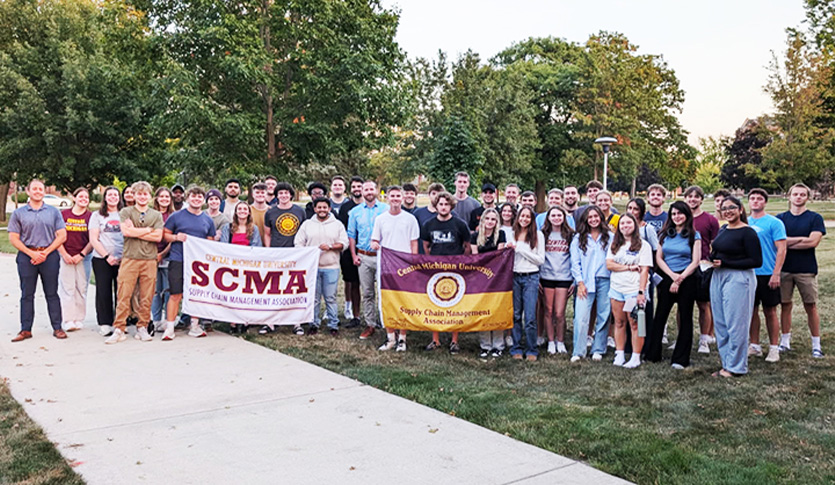 A large group poses outdoors on CMU’s campus, holding two banners for the Supply Chain Management Association.