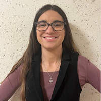 Jade Walleman smiling in a professional headshot, wearing glasses and a black vest against a textured wall.