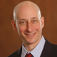 Spenser Robinson smiling in a professional headshot, wearing a suit and tie against a warm-toned background.