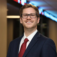 Trevor Carnovsky smiling in a professional headshot, wearing glasses and a suit with a red tie in an indoor setting.