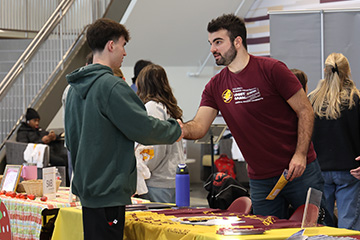 A student in a green hoodie and black pants shakes hands across a table with swag and brochures with a logistics department representative wearing a maroon and gold t-shirt at the CBA Spring Involvement Fair.