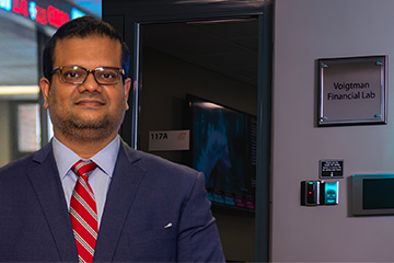 Abu Amin stands outside the Voigtman Financial Lab at Central Michigan University. He wears a blue suit, light blue shirt and red striped tie. Financial news displays are visible in the background.