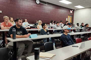 A group of twenty students sit at long tables in a CMU classroom. The students are facing forward and focused on the front of the classroom, which isn't visible in the image. The back wall of the class is painted maroon and the other side is white.