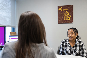 A student with silver hoop earrings and a black and white plaid flannel shirt sits at a desk facing an advisor who has her back to the camera.