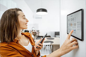 A woman stands in her dining room facing a agentic AI smart home screen on the wall. She is holding a phone at the same time as she points at icons on the wall-mounted screen.
