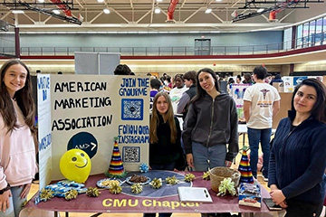 Four CMU students stand behind an American Marketing Association table at a campus organization event. AMA signage, QR codes, decorations and snacks are displayed. Sarah Firchau is on the far left, and Aimen Khan is on the far right.